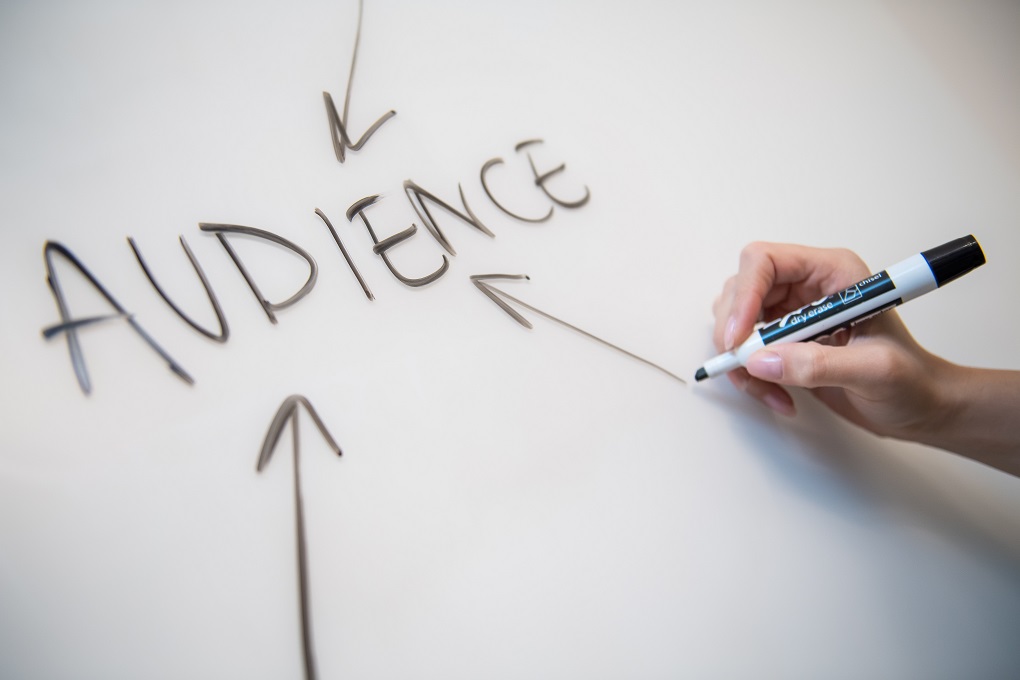 A hand holding a pen to write the word "AUDIENCE" on whiteboard